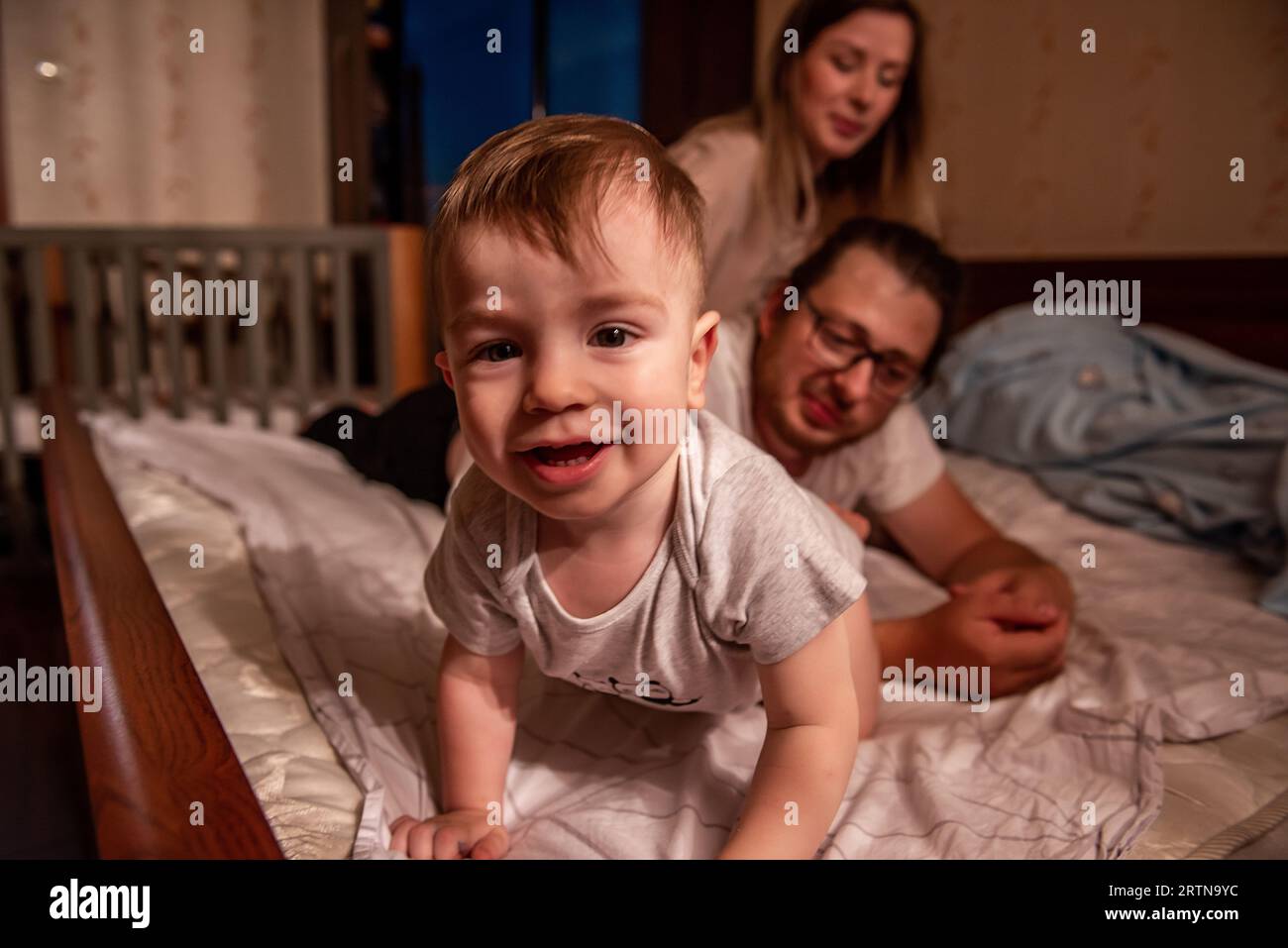 Baby boy lies on stomach on bed, smiling at camera. Family out of focus from behind. Out of focus, father and mother are laughing from behind, looking Stock Photo
