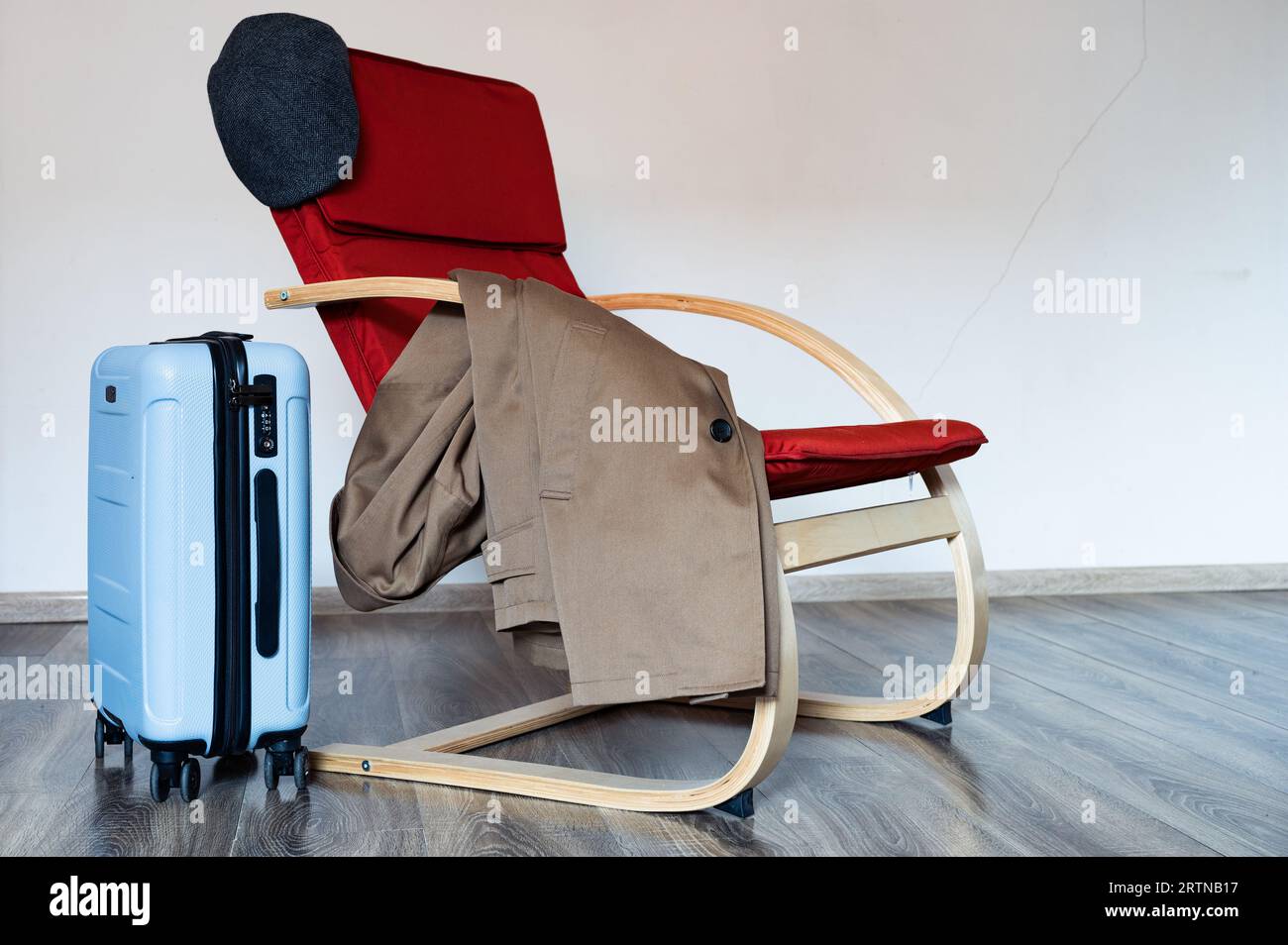 Travel blue suitcase stands near red armchair with coat and picky blinders hat on it Stock Photo