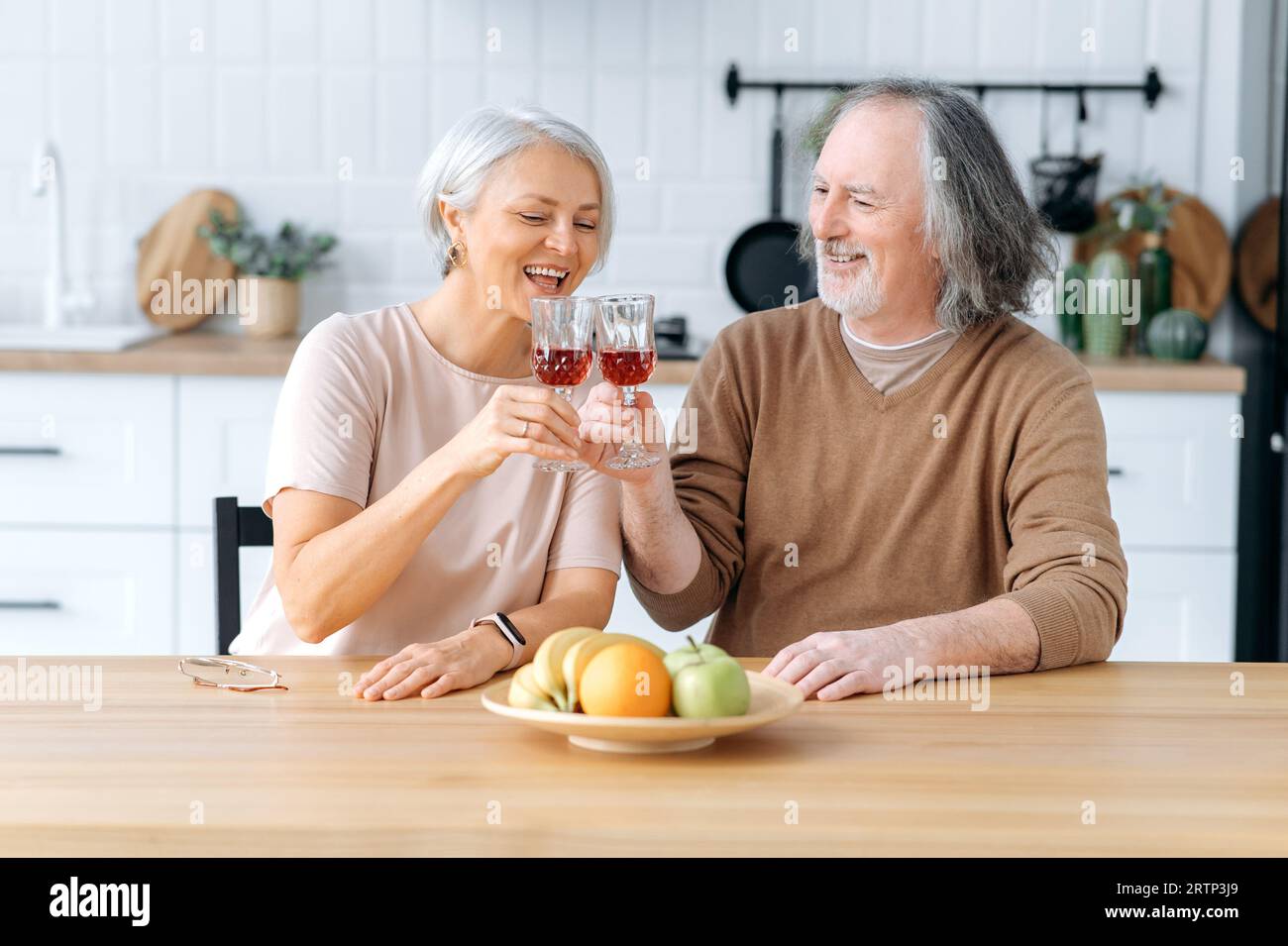 Cheers. Happy pensioners. Elderly caucasian spouses, sit at home in the kitchen, hold glasses of red wine in their hands, celebrate an important date, anniversary, look at glasses, smile joyfully Stock Photo