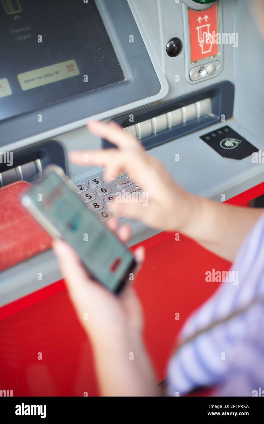 Unrecognizable woman holding a cell phone in front of a bank ATM carrying out an online banking operation. Stock Photo