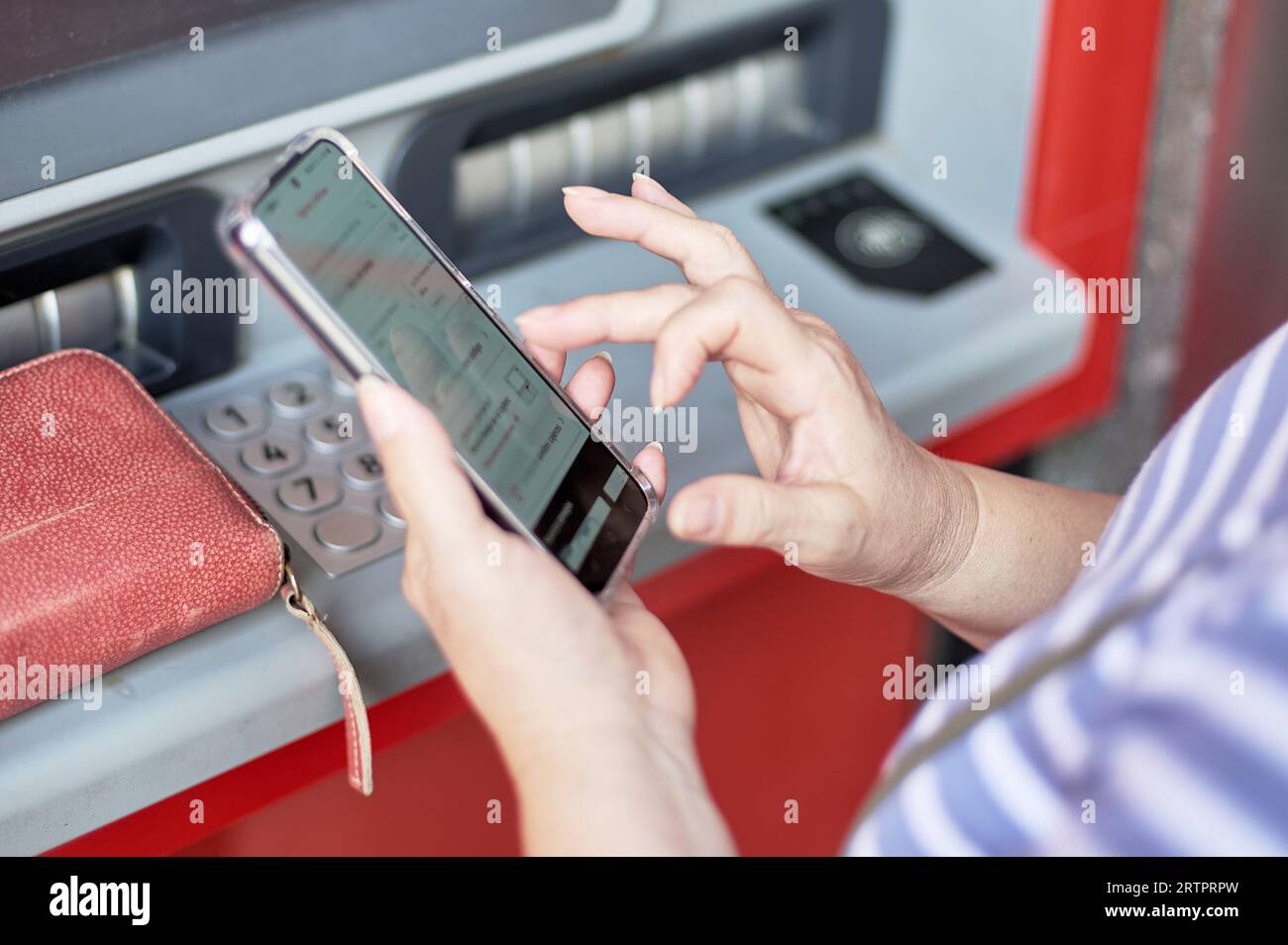 Unrecognizable woman holding a cell phone in front of a bank ATM carrying out an online banking operation. Stock Photo