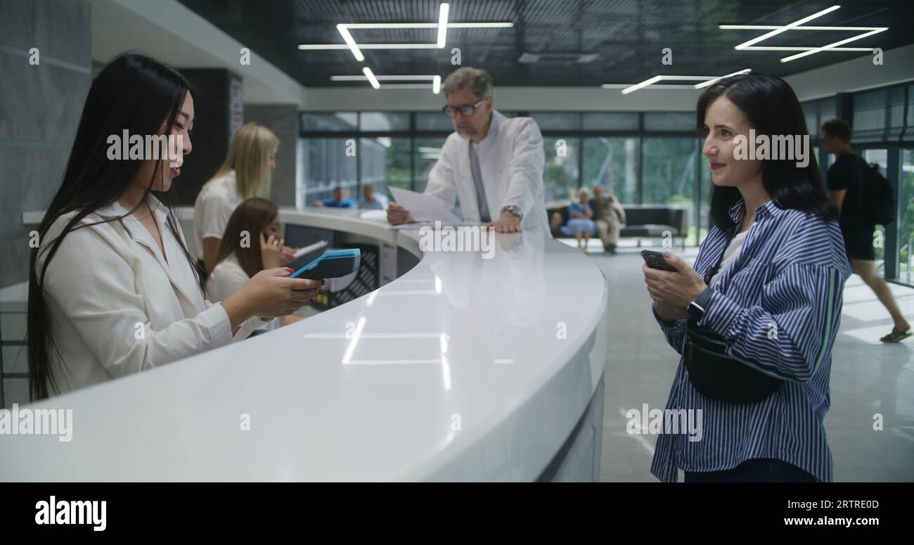 Asian female administrator at reception desk in clinic gives terminal ...