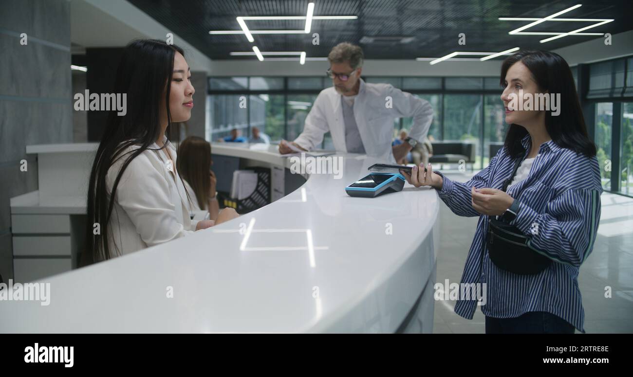 Asian female administrator at reception desk in clinic gives terminal ...