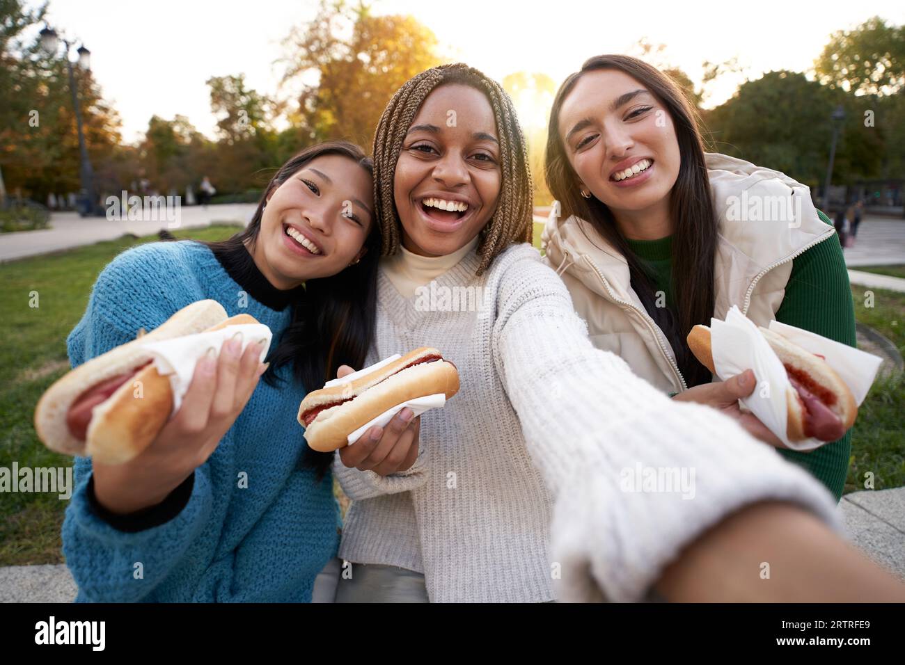 Group cheerful girls take selfie eating takeaway street food sitting on bench in nice area of city. Stock Photo