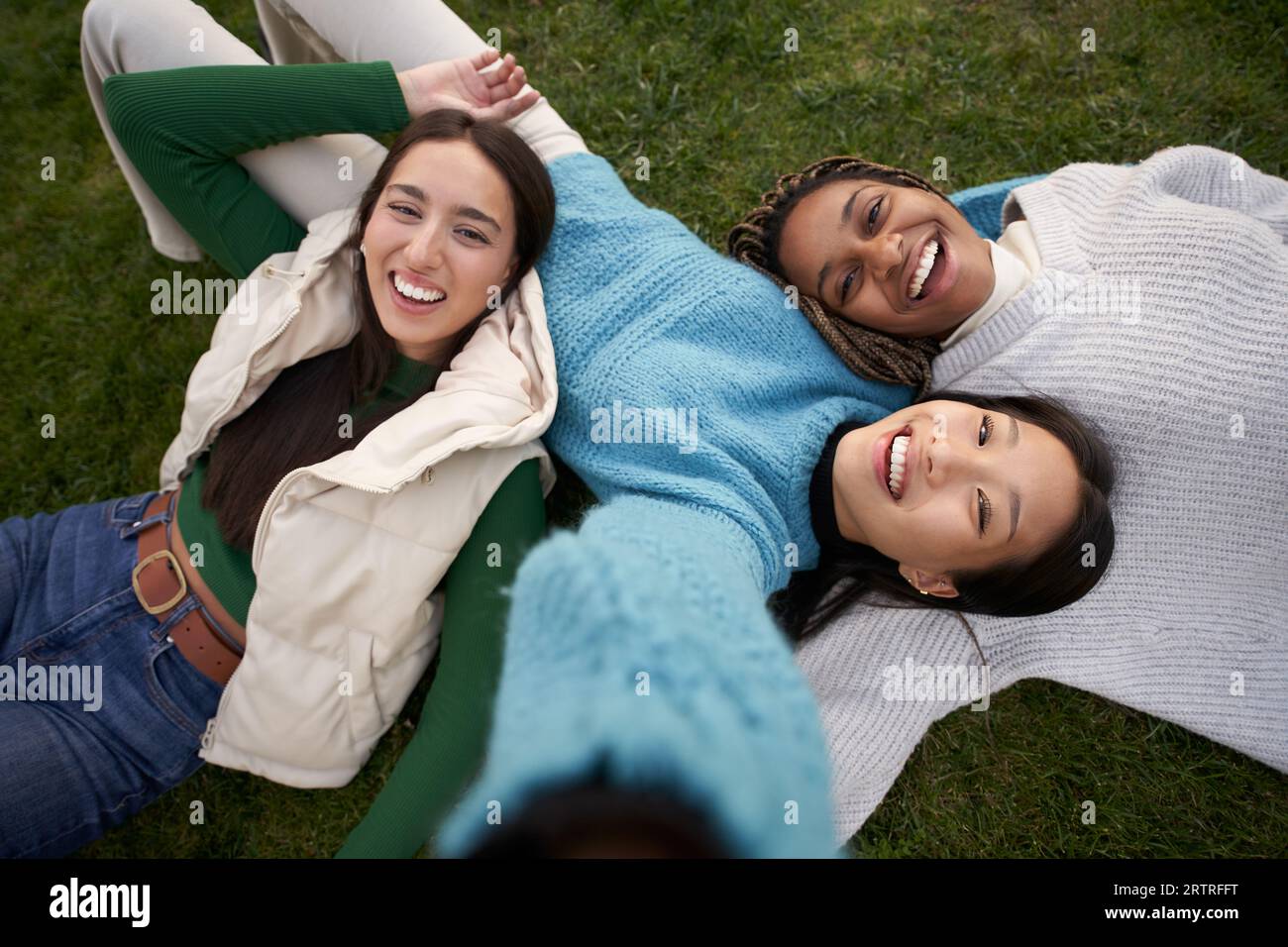 Three young multiracial girls lying on grass taking a selfie with cell phone looking at camera.  Stock Photo