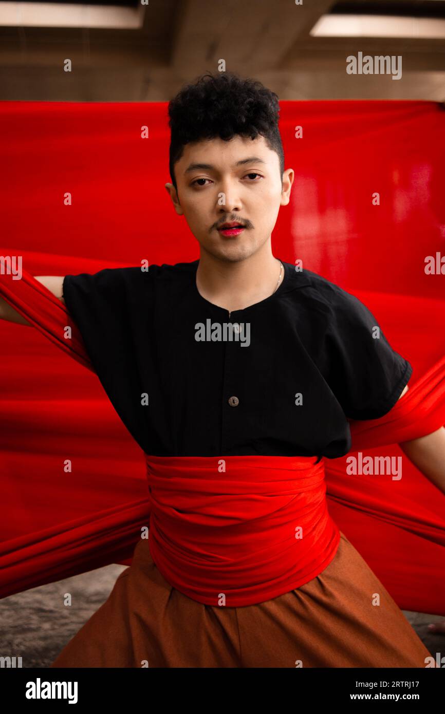 an Asian man holding a red cloth in his arms with a bold expression against a red background during the day Stock Photo