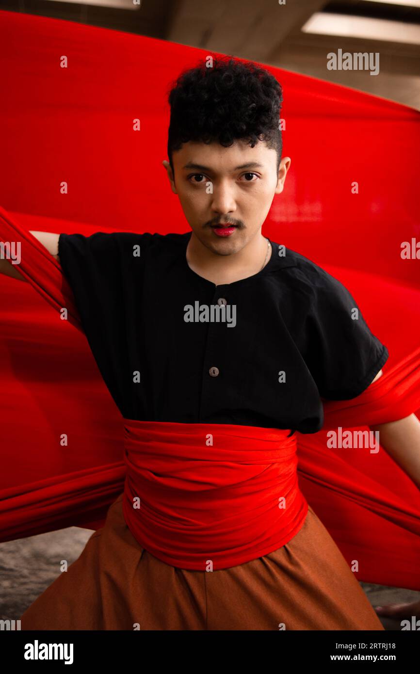 an Asian man holding a red cloth in his arms with a bold expression against a red background during the day Stock Photo