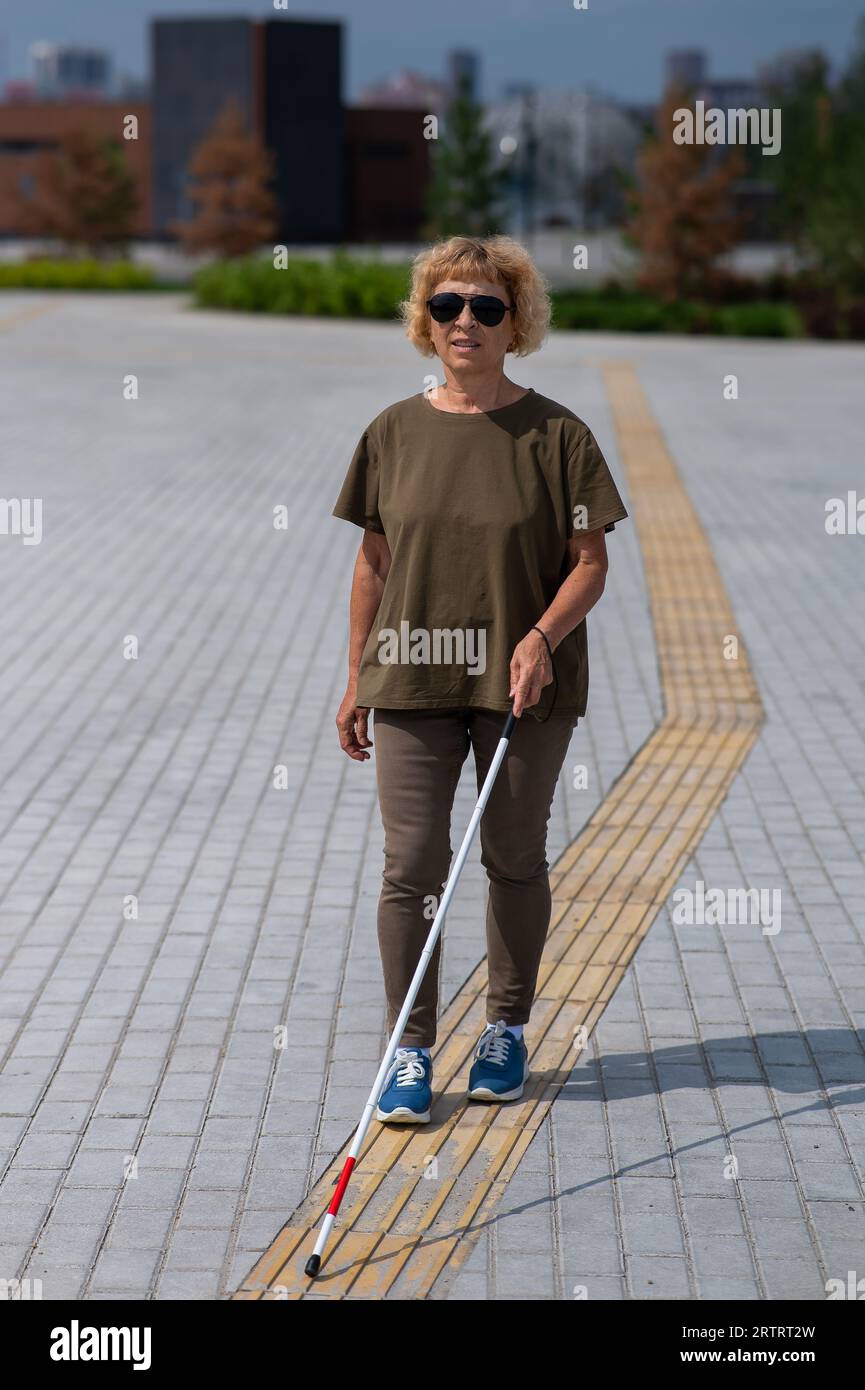 An elderly blind woman walks with a cane along a tactile tile Stock