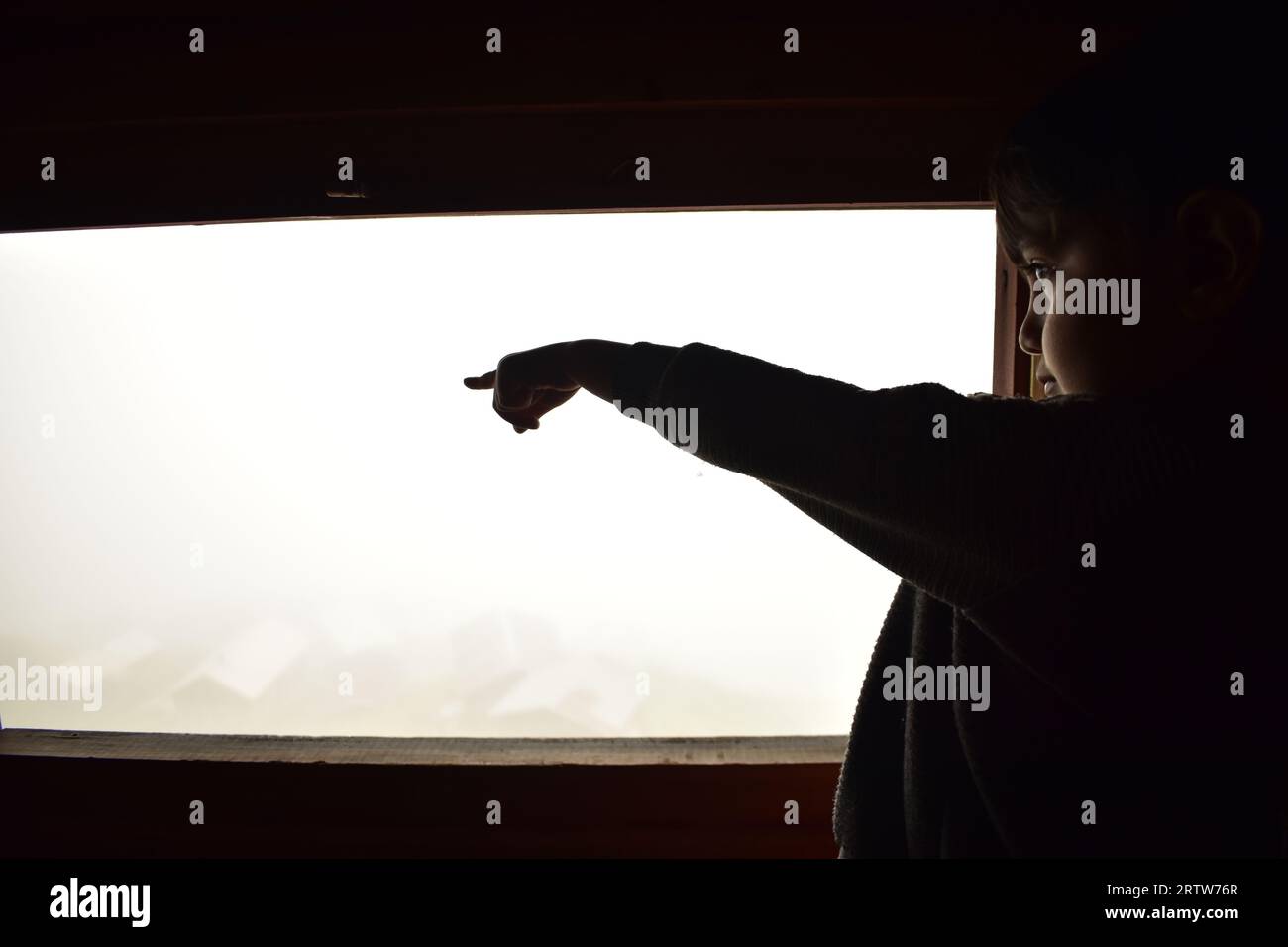 A happy kid pointing outside from a window of a mountain house in a foggy morning Stock Photo