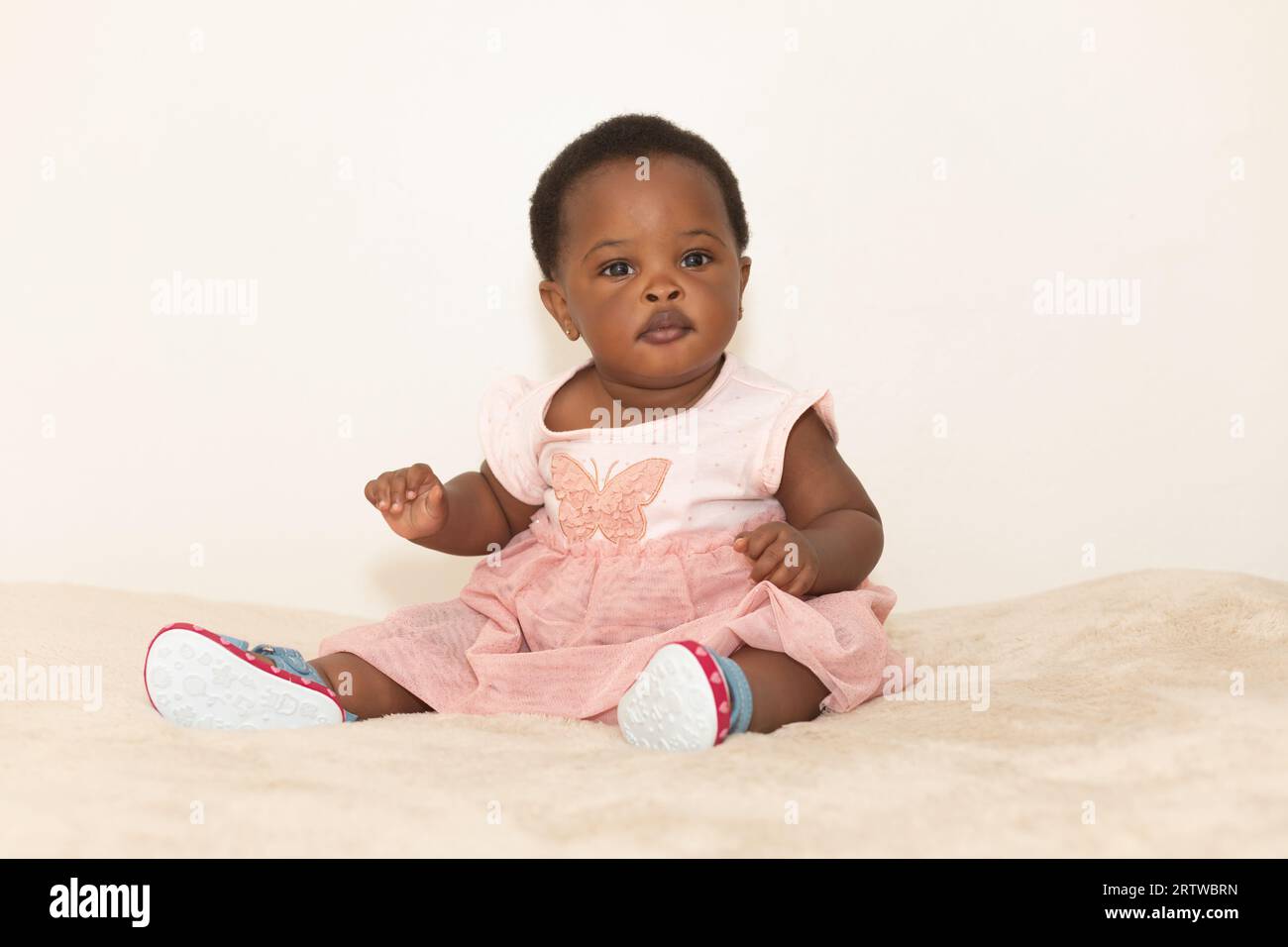 Portrait of a black baby girl dressed in a pink dress sitting on a bed Stock Photo