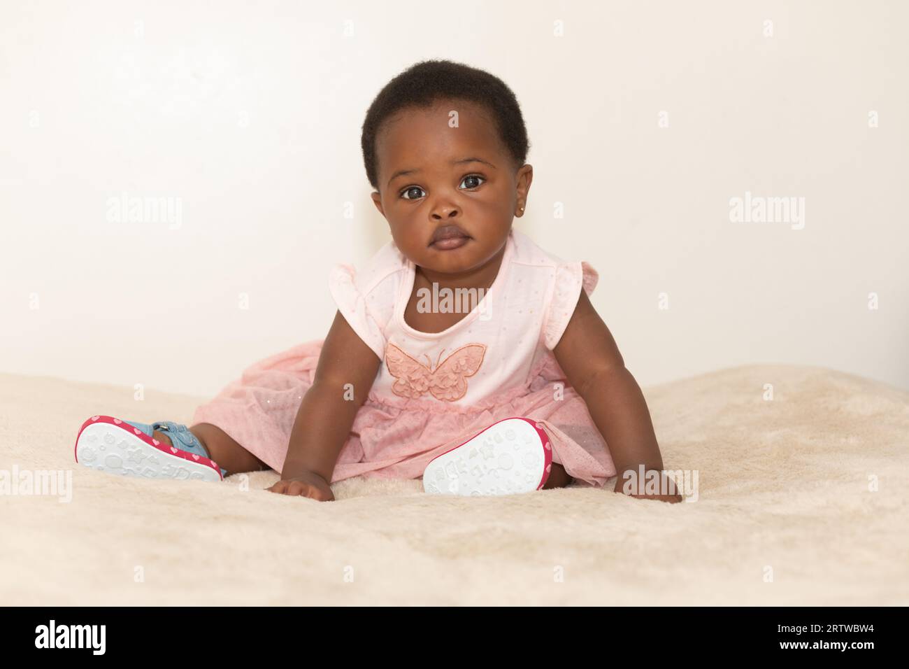 Portrait of a black baby girl dressed in a pink dress sitting on a bed Stock Photo