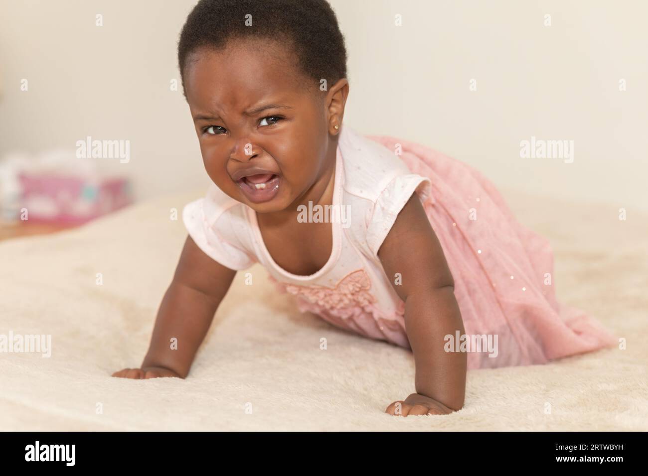 Portrait of a crying black baby girl dressed in a pink dress crawling on a bed Stock Photo