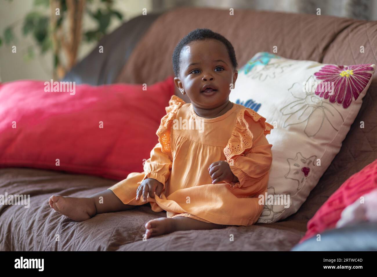 Portrait of a smiling black baby girl dressed in an orange dress sitting on a couch Stock Photo