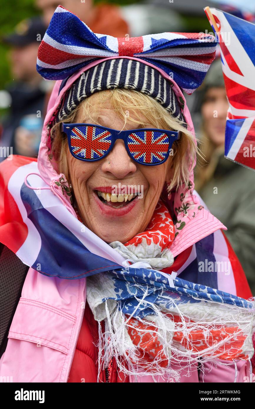 A woman wearing Union Jack glasses is part crowd braving the rain to watch the coronation of King Charles III, on a giant TV screen. Hyde Park, London Stock Photo