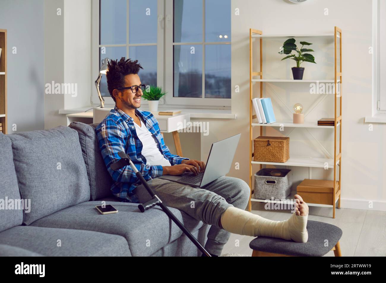 Young man with broken leg in plaster cast sitting on couch with laptop Stock Photo