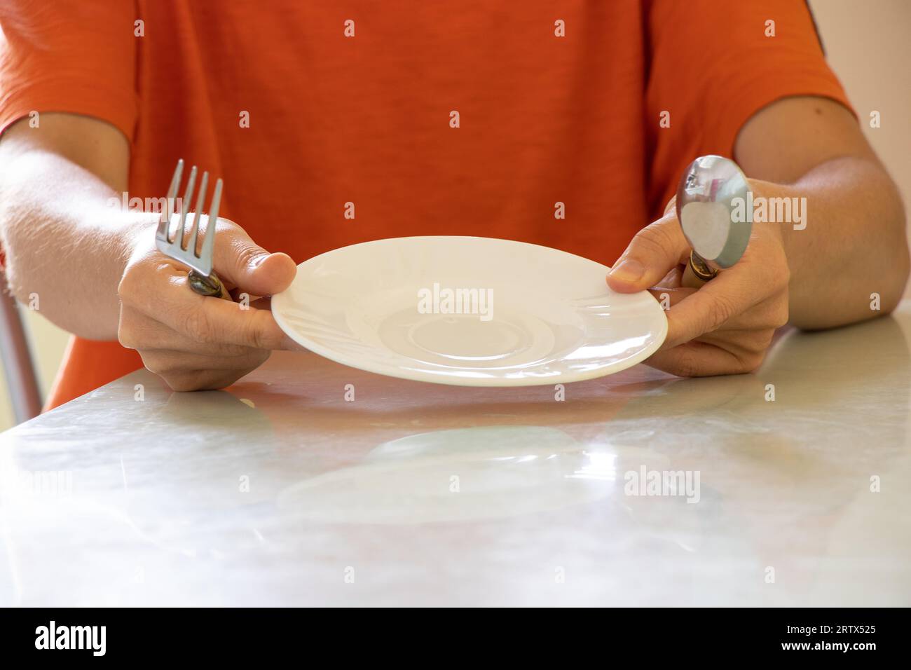 A girl with a spoon and fork and an empty plate sits at the table in the kitchen at home, without food, nothing to eat Stock Photo