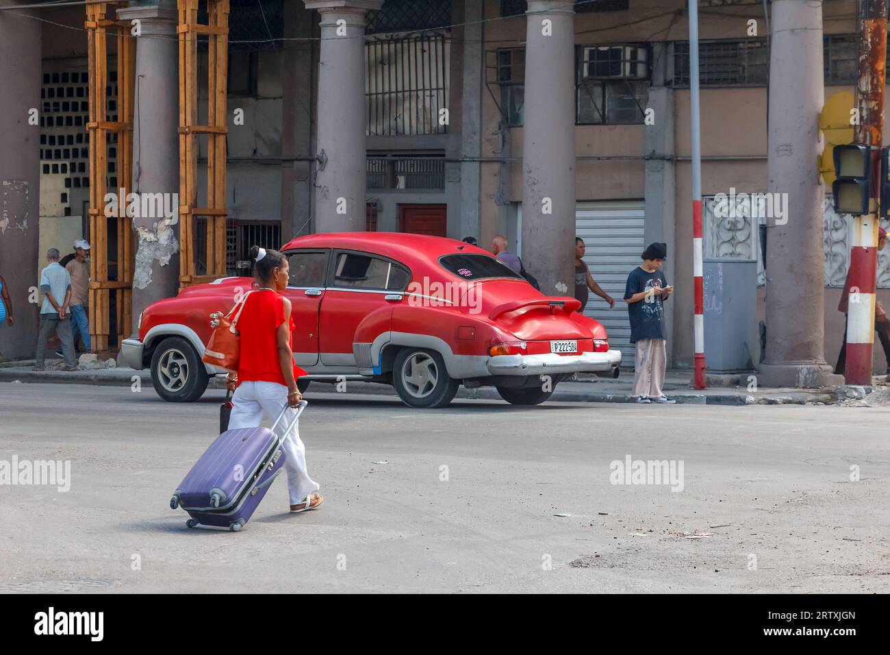 Havana, Cuba, 2023, A Cuban woman pulls a wheeled suitcase on a city street. A red vintage American car drives in the scene. Stock Photo