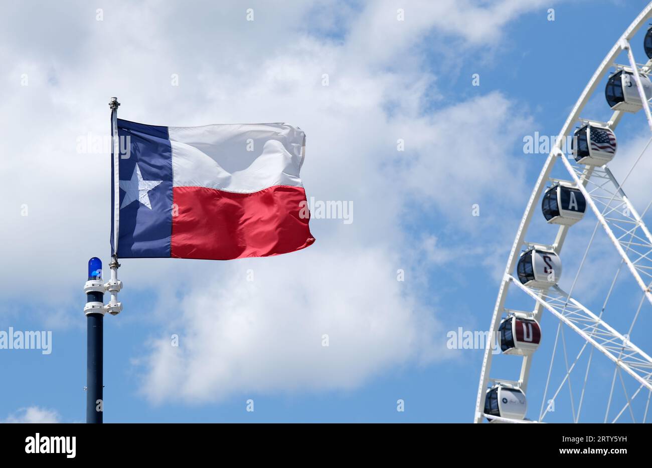 Texas state flag flying against a blue sky and white cloud background. Partial view of a Ferris wheel included. Stock Photo