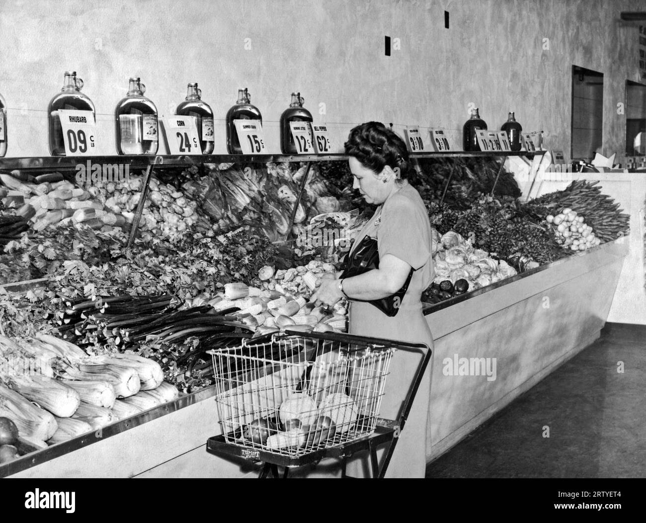 United States    c. 1940. A woman in a grocery store making selections from the vegetable department. She is using an early shopping cart that carries the separate basket and folds up when not in use. Stock Photo
