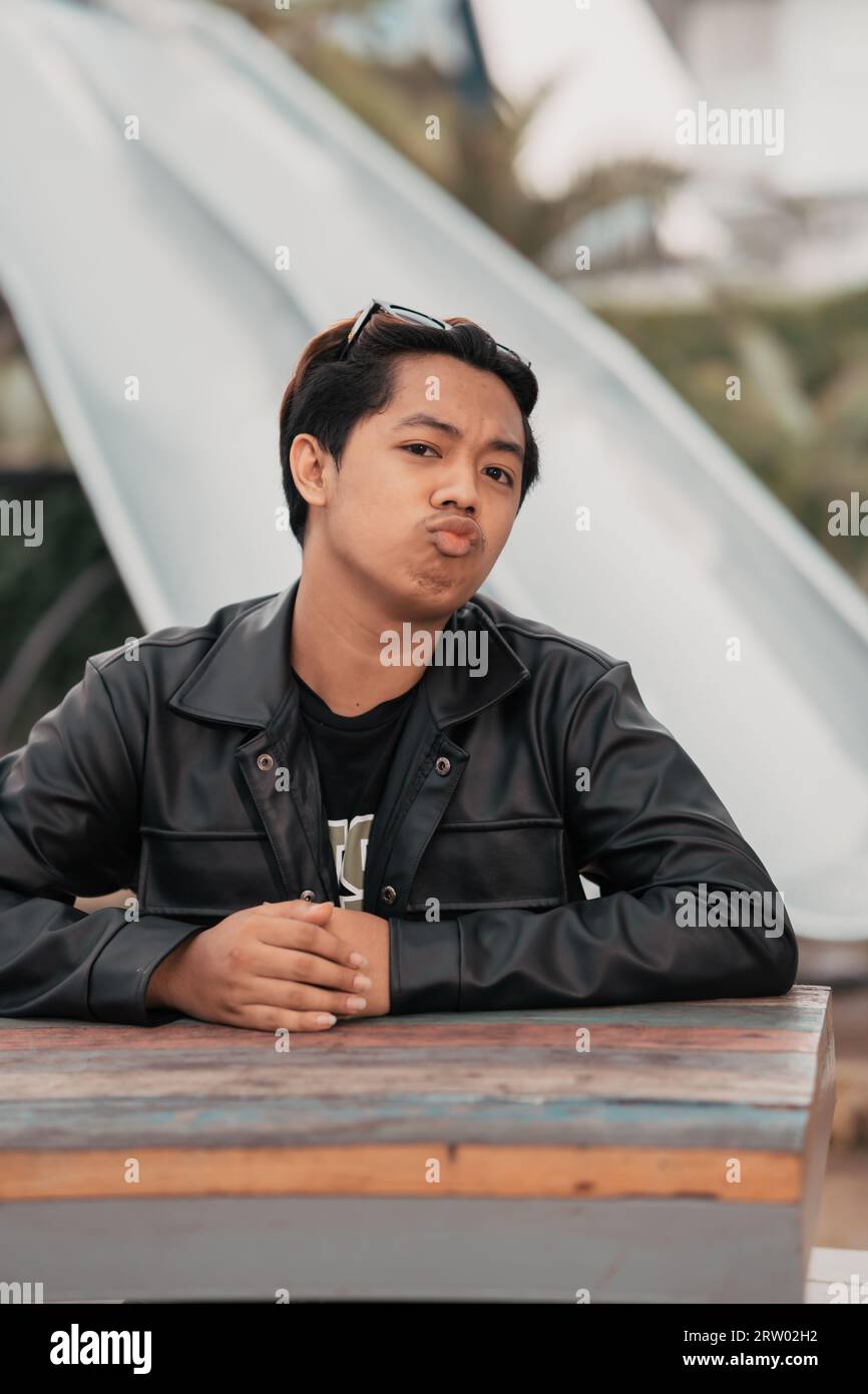 an Asian man with a chubby face wearing sunglasses and a black leather jacket while sitting at a cafe table in the morning Stock Photo