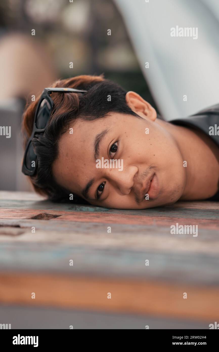 an Asian man with a chubby face wearing sunglasses and a black leather jacket while sitting at a cafe table in the morning Stock Photo