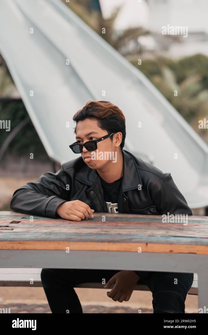 an Asian man with a chubby face wearing sunglasses and a black leather jacket while sitting at a cafe table in the morning Stock Photo