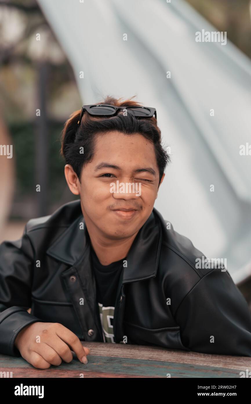 an Asian man with a chubby face wearing sunglasses and a black leather jacket while sitting at a cafe table in the morning Stock Photo