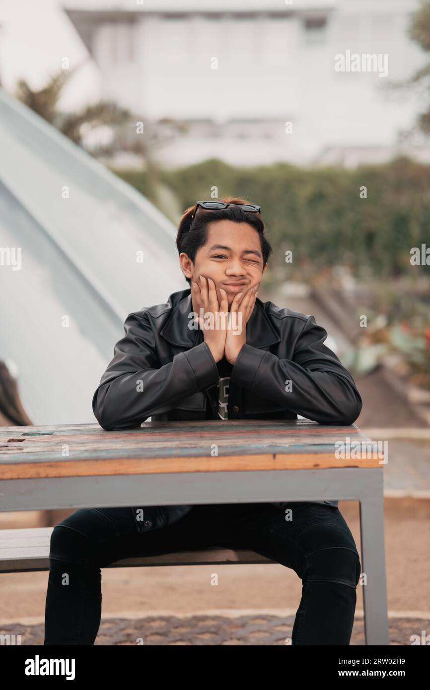 an Asian man with a chubby face wearing sunglasses and a black leather jacket while sitting at a cafe table in the morning Stock Photo