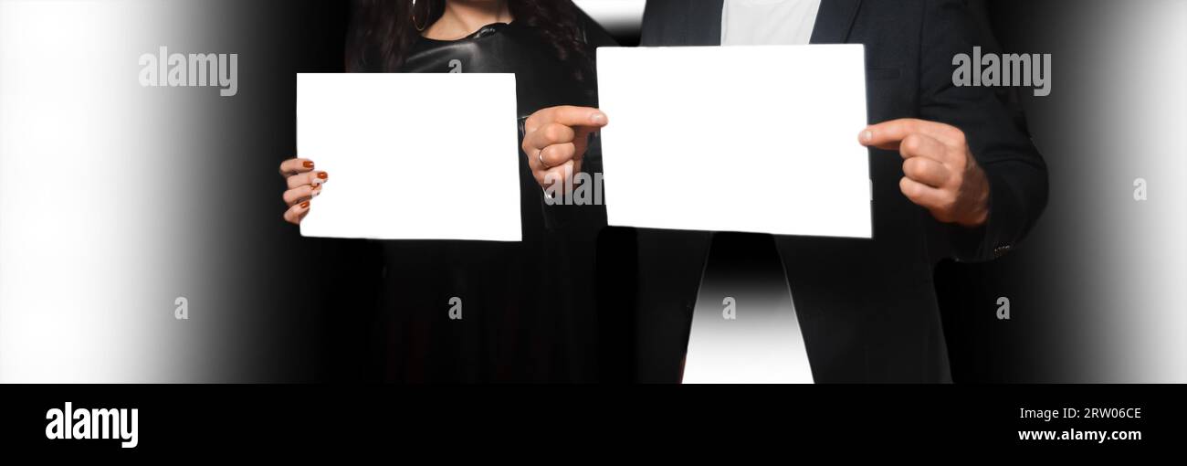 Two people, a man and a woman, holding a sheet of A4 paper in their hands, an empty space for text and design white blank on a dark background. Stock Photo
