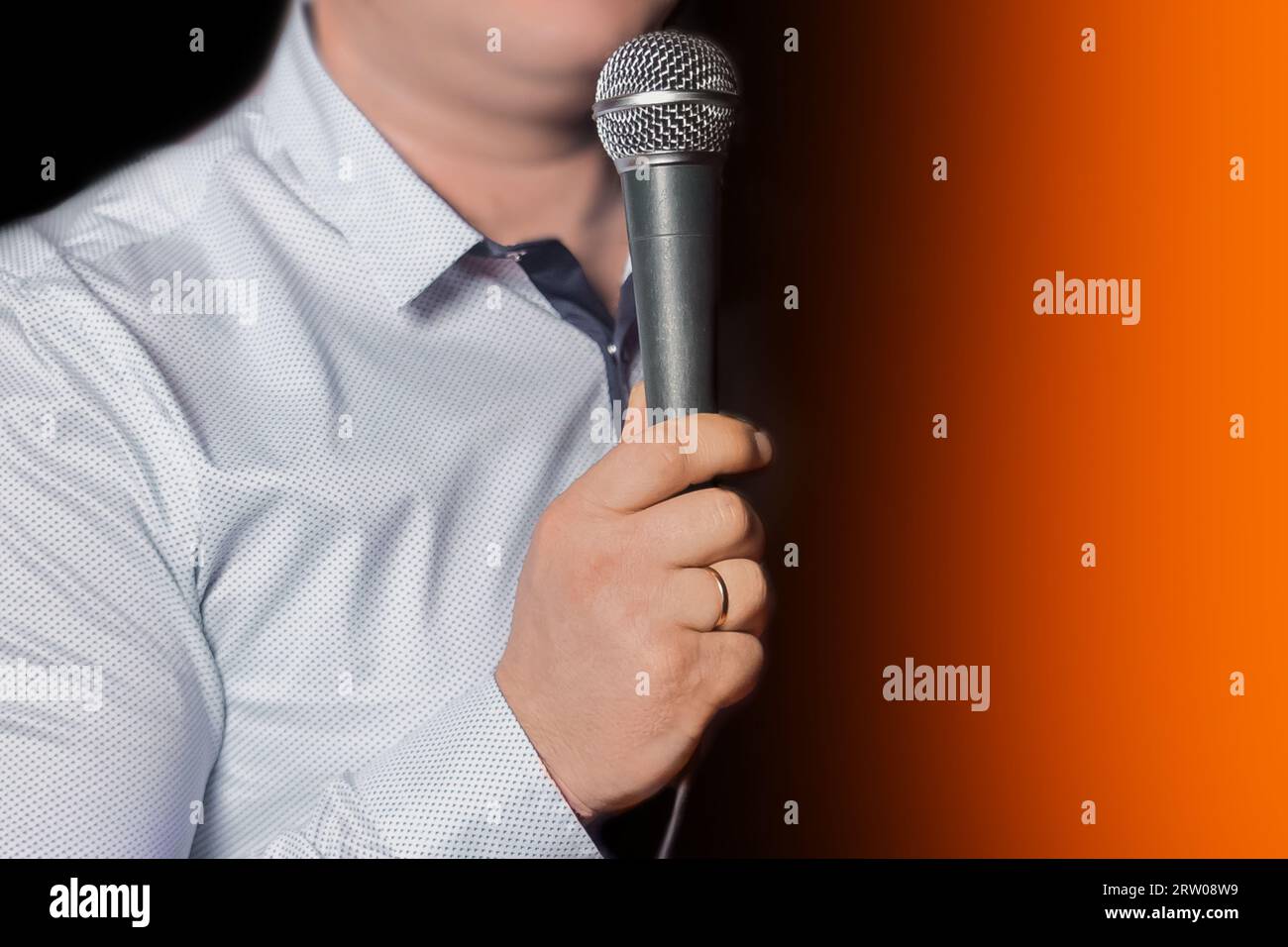 Person's hand with a ring on his finger holding a professional instrument karaoke equipment microphone for singing on a black orange background close- Stock Photo