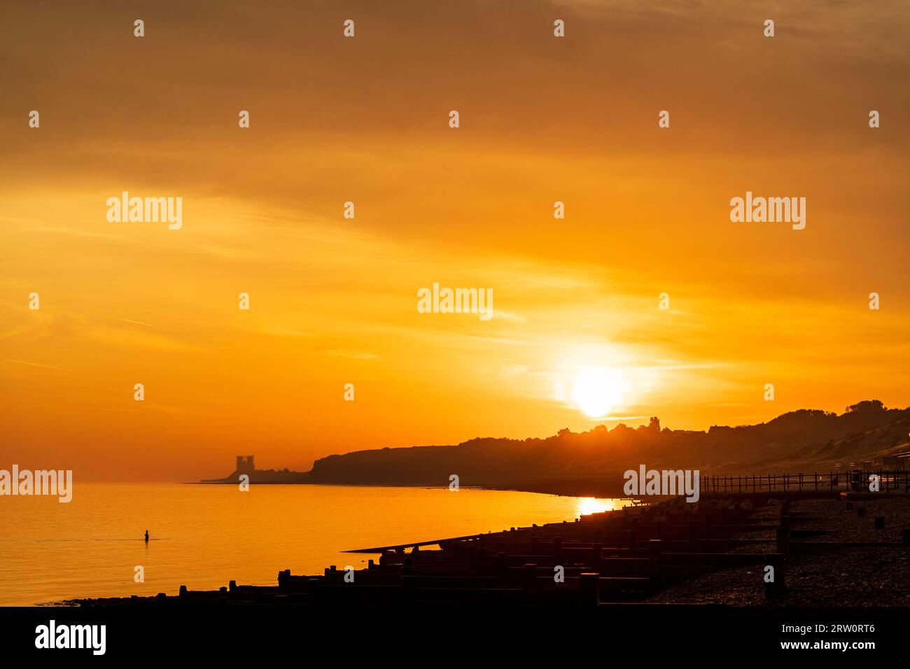 Sunrise at Herne Bay in Kent. Orange sky and sea with the bright sun appearing above silhouetted coastline. In the distance, the landmark twin towers of the ruins of the Angelo Saxon church at Reculver. Breakwaters along the beach, very calm sea. Stock Photo
