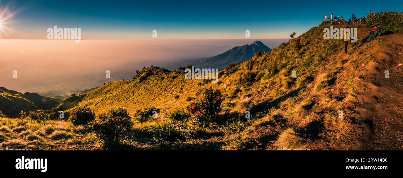 Panoramic photo of mountains surrounding Mount Merbabu and hikers at top during sunrise near Yogya in central Java province in Indonesia. In this Stock Photo