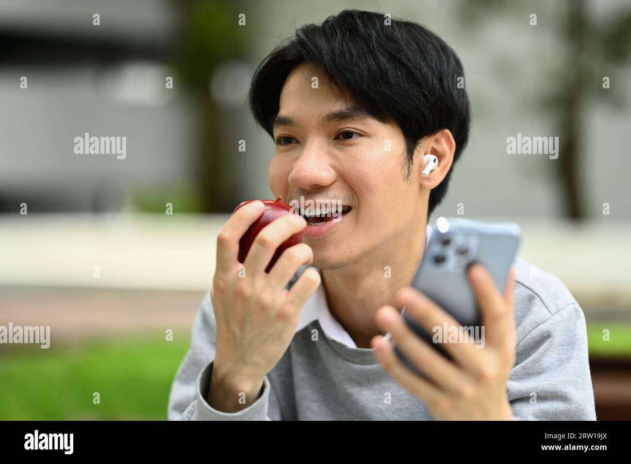 Smiling young asian man eating apple and using smart phone while ...