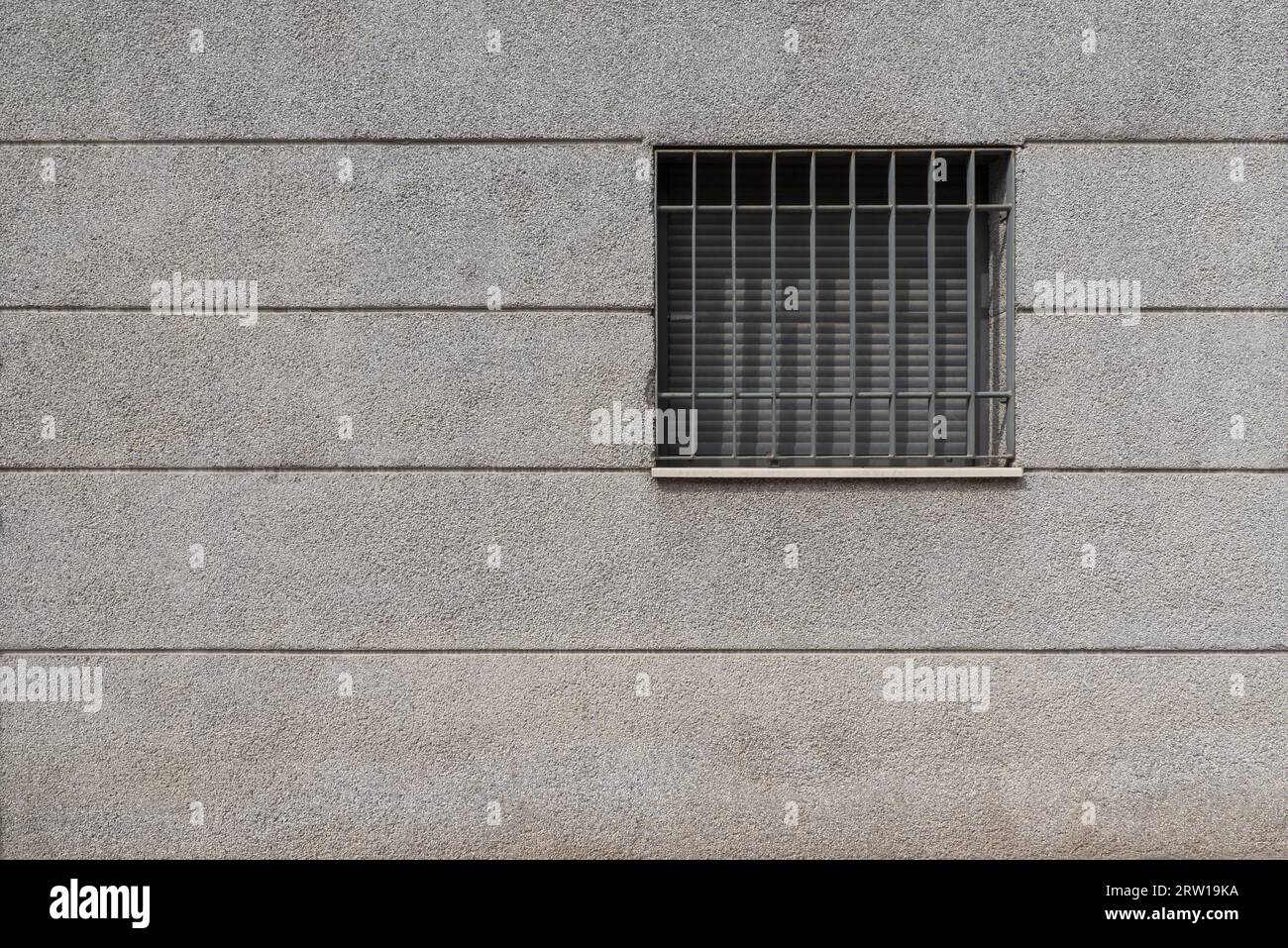 A square window with the blinds lowered and a metal grill of the same color on a wall plastered with cement and pebbles Stock Photo
