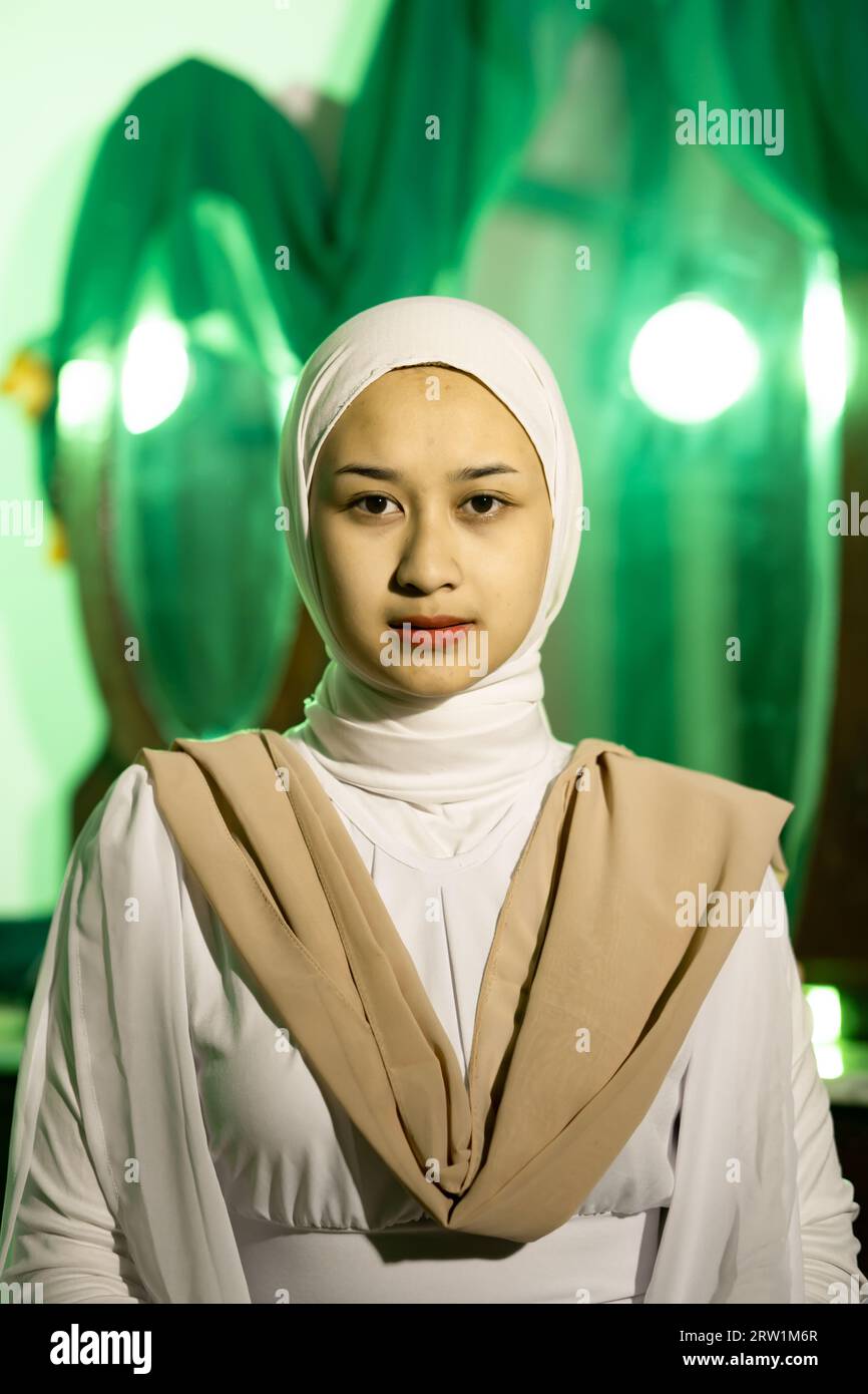 a Muslim woman with a white headscarf and white clothes is sitting in the middle of a green room