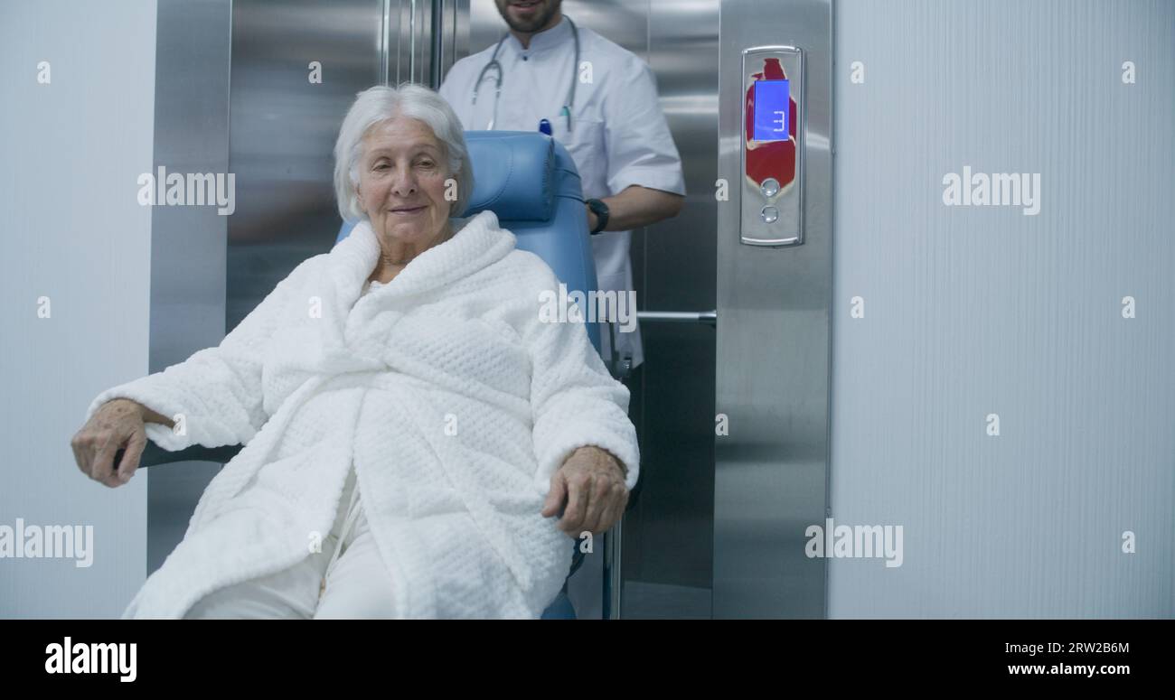 Doctor moves elderly woman in transport wheelchair from elevator. Medic ...
