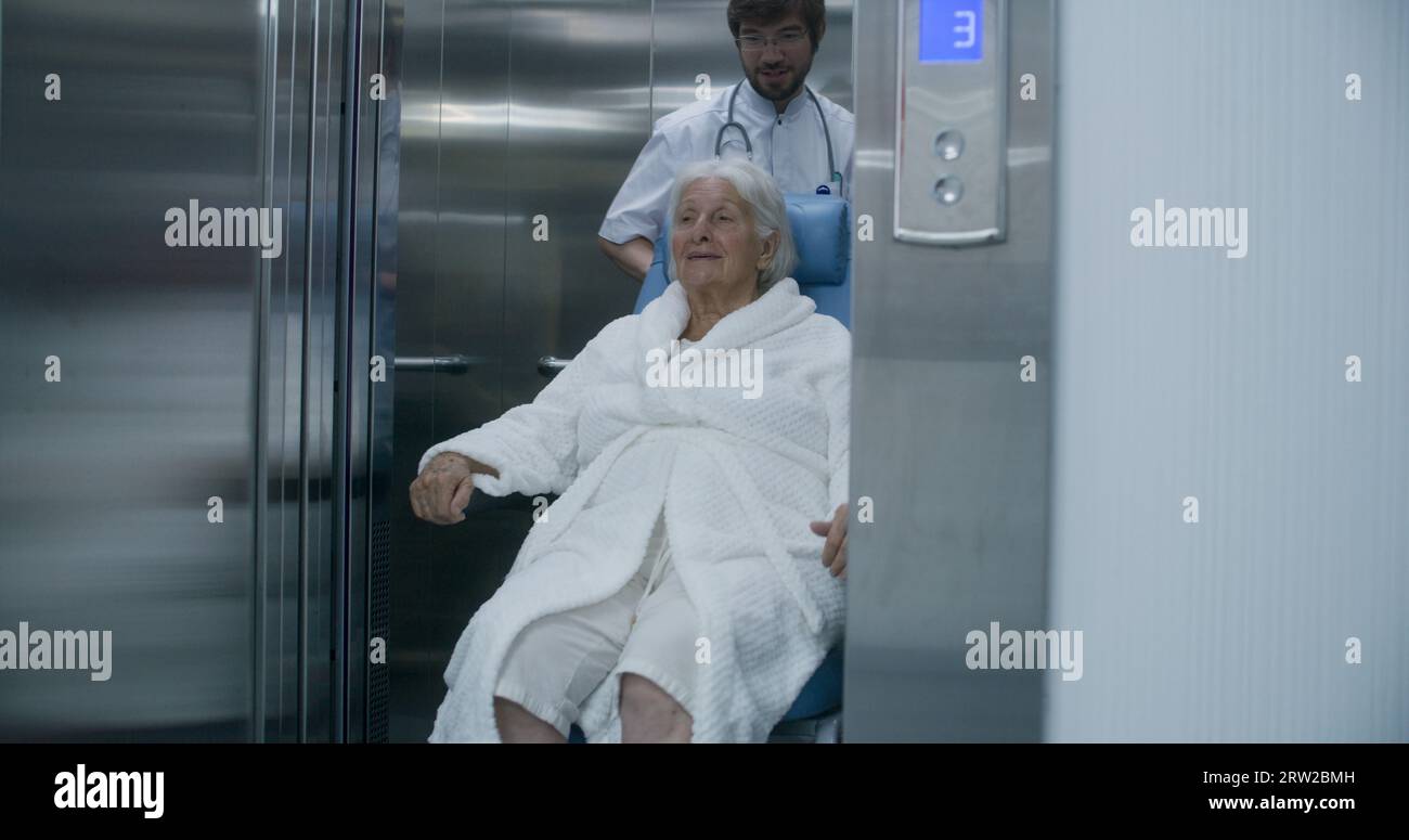 Doctor moves elderly woman in transport wheelchair from elevator. Medic ...