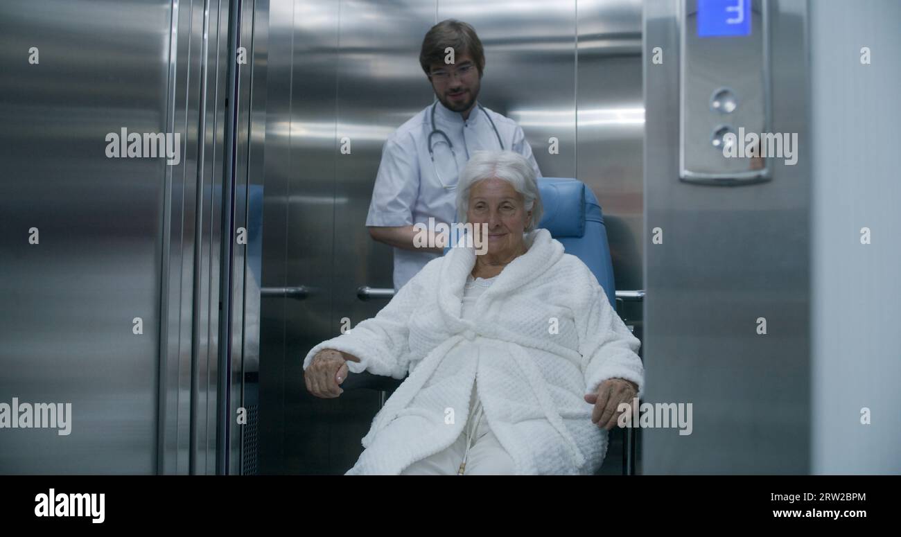 Doctor moves elderly woman in transport wheelchair from elevator. Medic ...