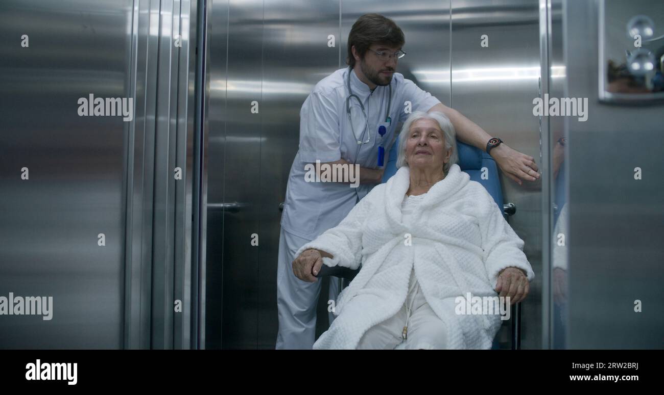 Doctor moves elderly woman in transport wheelchair from elevator. Medic ...