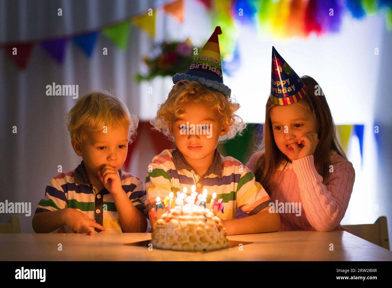 Kids birthday party. Children blow out candles on cake in dark room. Rainbow decoration and table setting for kids event, banner and flag. Stock Photo