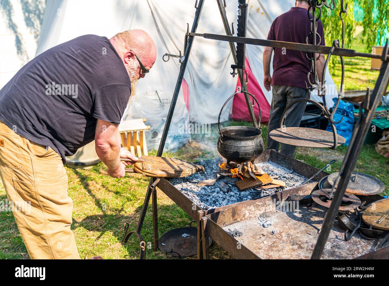 Middle aged man using medieval bellows on a small fire of chopped wood with a cauldron hanging over on a cast iron frame work. Stock Photo