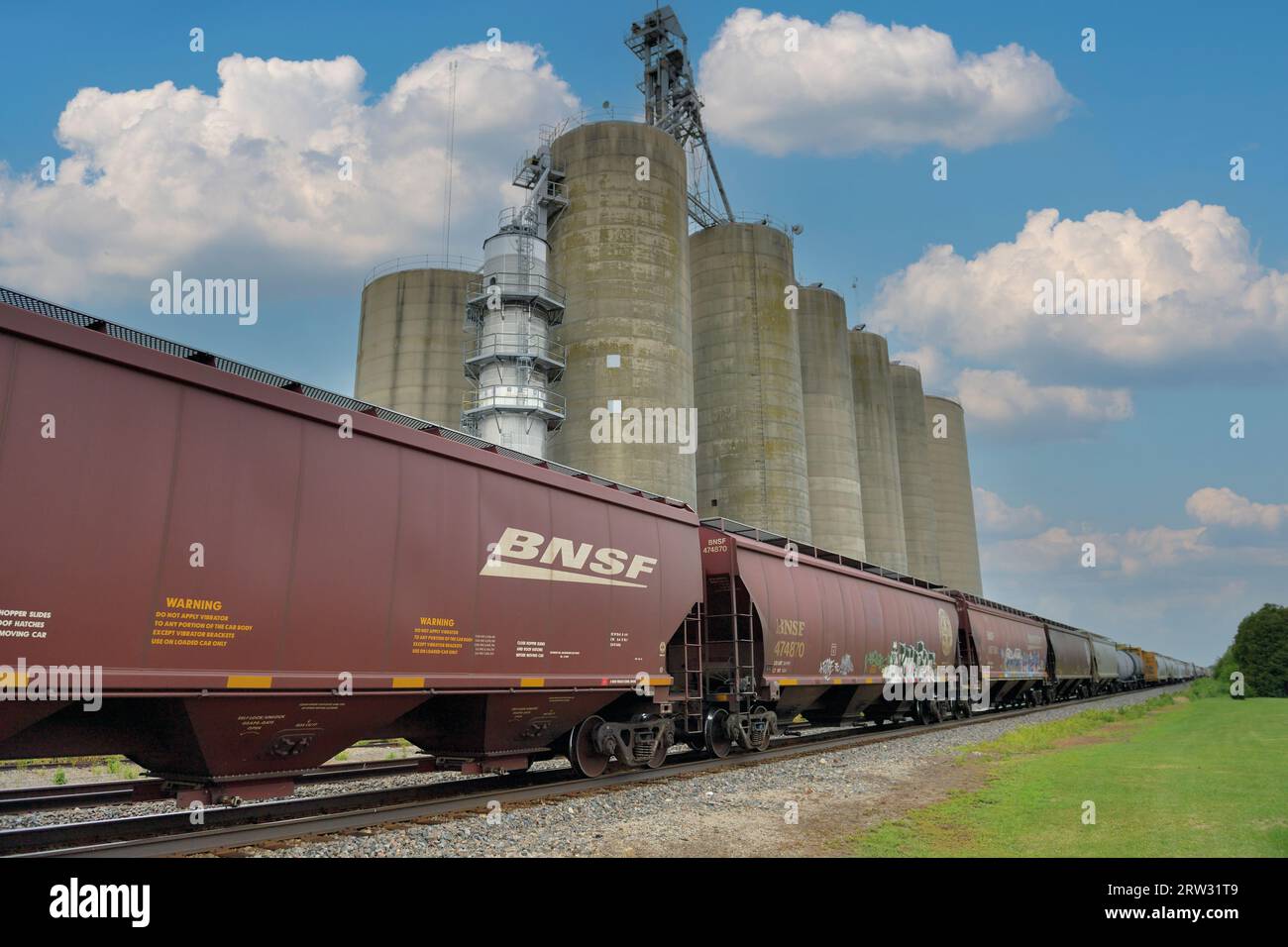 Chana, Illinois, USA. A Burlington Northern Santa Fe grain train roars by a large grain elevator while traveling through north central Illinois. Stock Photo