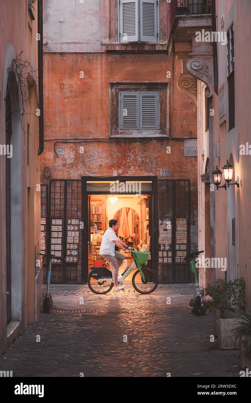 Rome, Italy - August, 27th, 2023: A man on a hired bicycle cycles over ancient cobblestone streets in front of an Italian leather shop at night in the Stock Photo