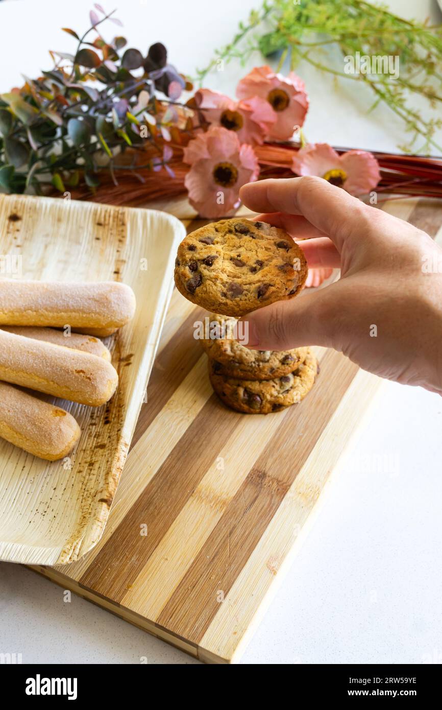 Woman's hand holding a homemade cookie with chocolate chips from a decorated table. Vertical shot. Stock Photo