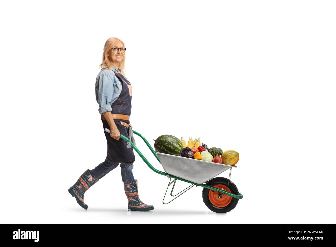 Full length profile shot of a woman farmer pushing a wheelbarrow full of fruits and vegetables isolated on white background Stock Photo