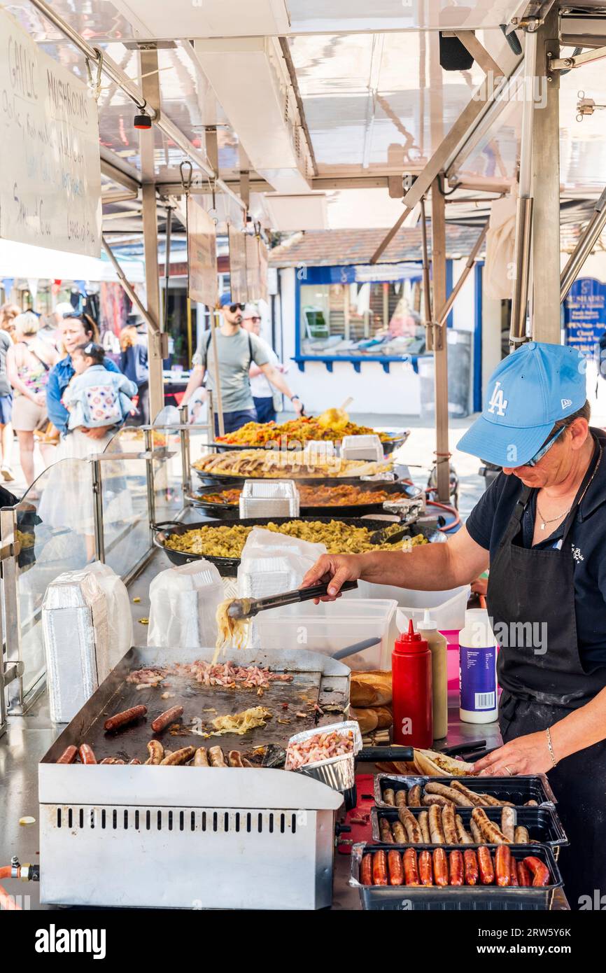 French food stall in the market square in the Kent town of Sandwich. A Woman preparing sausages for hot dogs on hot plate while people wait. Stock Photo