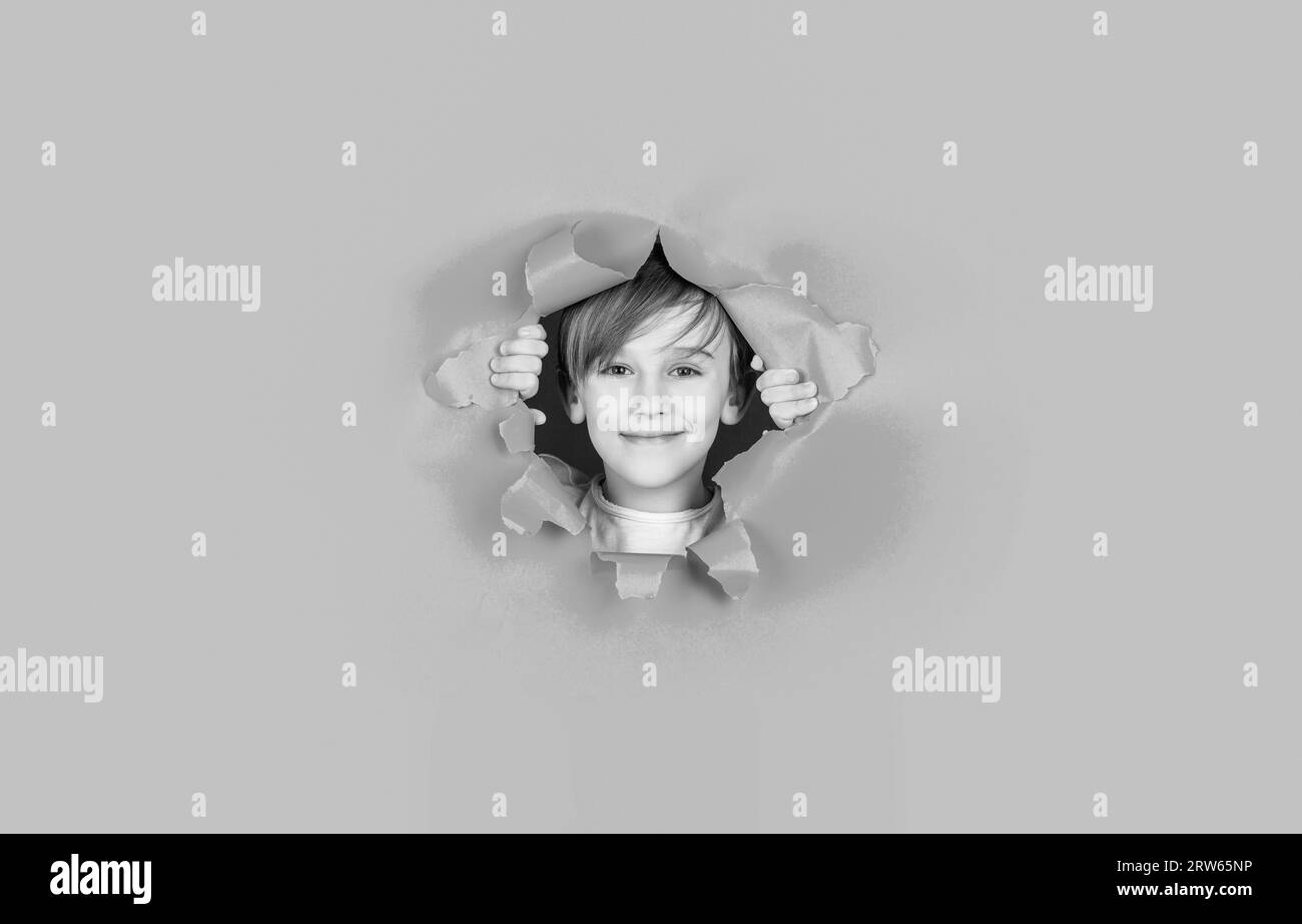 Kid with toothy smile shows face in paper hole. Positive child with toothy pleasant smile on face, keeps through torn hole in paper. Black and white Stock Photo