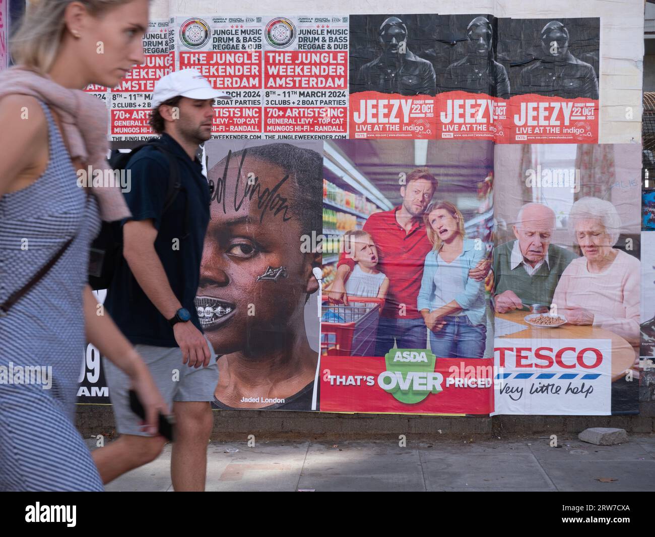 Cost of Living crisis UK. People walking past a protest advert for Tesco and Asda, demonstrating the high profits  supermarkets are making during the cost of living crisis. Type on spoof adverts read Tesco very little help, and Asda that's over priced Stock Photo