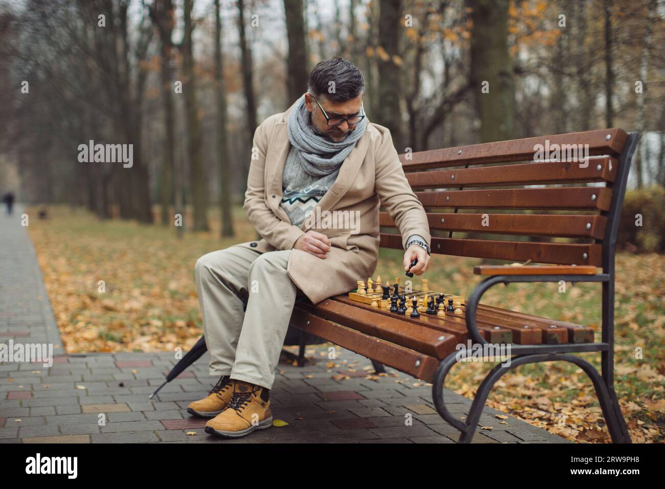 An elderly man plays chess on a bench in an autumn park. Stock Photo