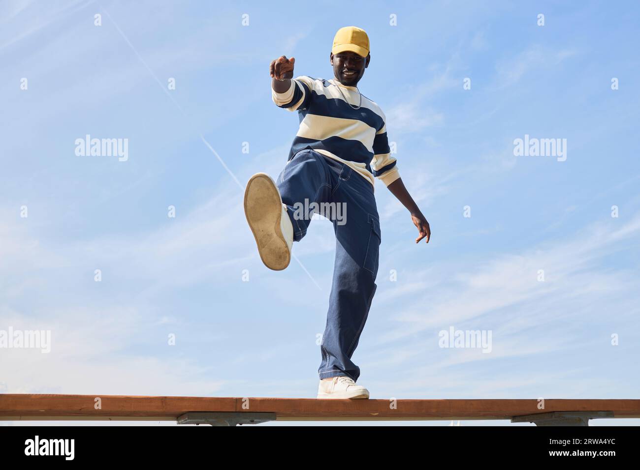 Minimal fashion shot of young black man wearing trendy outfit standing on roof against blue sky and kicking off to camera. Stock Photo